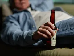 Mężczyzna w dżinsach i koszuli trzyma butelkę piwa, leżąc na kanapie. Może to sugerować spalanie alkoholu podczas snu.