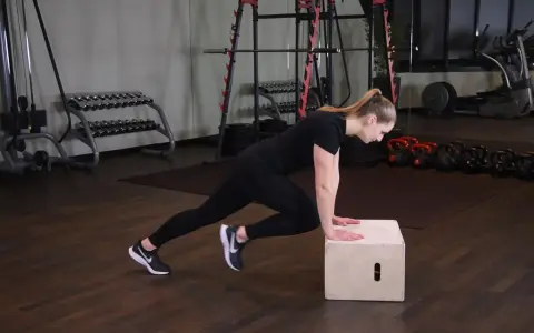 Kobieta wykonuje ćwiczenie mountain climbers na skrzyni. W tle siłownia z ciężarami.