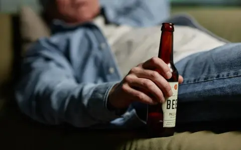 Mężczyzna w dżinsach i koszuli trzyma butelkę piwa, leżąc na kanapie. Może to sugerować spalanie alkoholu podczas snu.