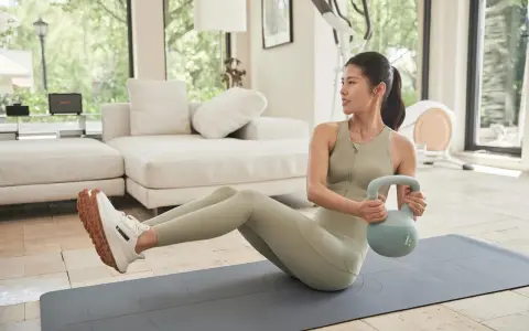 Kobieta wykonuje ćwiczenia na brzuch z kettlebell, siedząc na macie w domowej siłowni.
