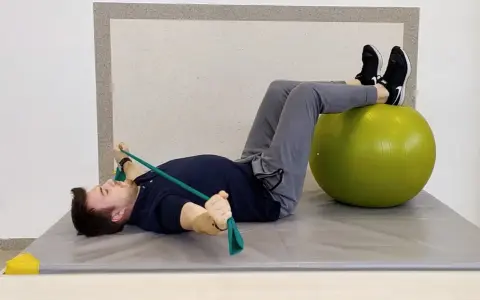Mężczyzna ćwiczy z taśmą oporową i piłką gimnastyczną, leżąc na macie.