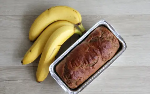 Gotowy proteinowy chlebek bananowy w formie, obok pęk dojrzałych bananów.