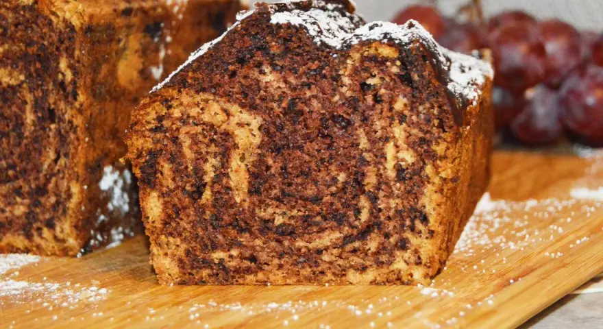 Puszysty chlebek bananowy z kakao, oprószony cukrem pudrem, prezentuje się apetycznie na drewnianej desce.
