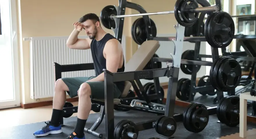Młody mężczyzna, pierwszy raz na siłowni, siedzi zmęczony na ławce, wycierając pot z czoła. Wokół niego sprzęt do ćwiczeń.