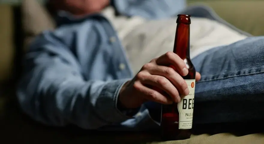 Mężczyzna w dżinsach i koszuli trzyma butelkę piwa, leżąc na kanapie. Może to sugerować spalanie alkoholu podczas snu.