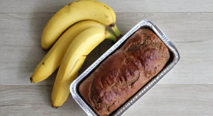 Gotowy proteinowy chlebek bananowy w formie, obok pęk dojrzałych bananów.