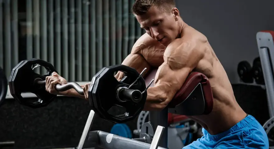 Mężczyzna wykonuje ćwiczenia na biceps i triceps z użyciem sztangi łamanej na modlitewniku.