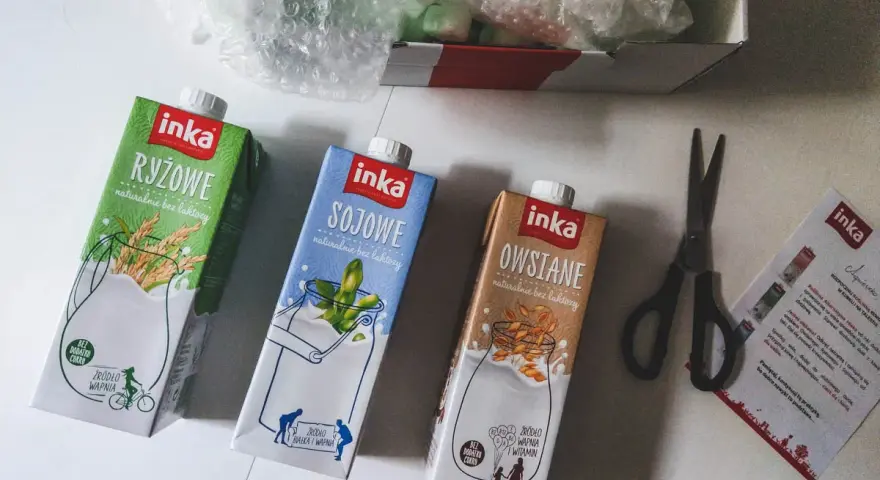 Trzy kartony napojów roślinnych Inka: ryżowy, sojowy i owsiany. Obok nożyczki i ulotka. Tak powstaje mleko owsiane.