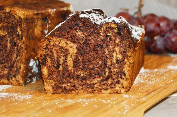 Puszysty chlebek bananowy z kakao, oprószony cukrem pudrem, prezentuje się apetycznie na drewnianej desce.